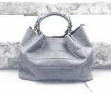 A woven braided pattern black leather large handbag with a zippered top, detachable shoulder strap, and a textured surface, placed on a stone bench.