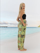 Model wearing green boho pants with geometric patterns, standing on a beach with the ocean in the background. The pants are loose and comfortable, featuring drawstrings at the waist and ankles for a relaxed fit.