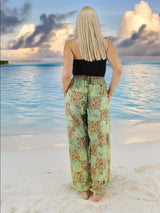 Model wearing green boho pants with geometric patterns, standing on a beach with the ocean in the background. The pants are loose and comfortable, featuring drawstrings at the waist and ankles for a relaxed fit.
