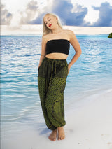 Model wearing green boho pants with geometric patterns, standing on a beach with the ocean in the background. The pants are loose and comfortable, featuring drawstrings at the waist and elastic at the ankles for a relaxed fit