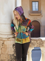 Colorful patchwork boho hoodie with multicolored stripes, worn by a young female model against a stone wall backdrop.